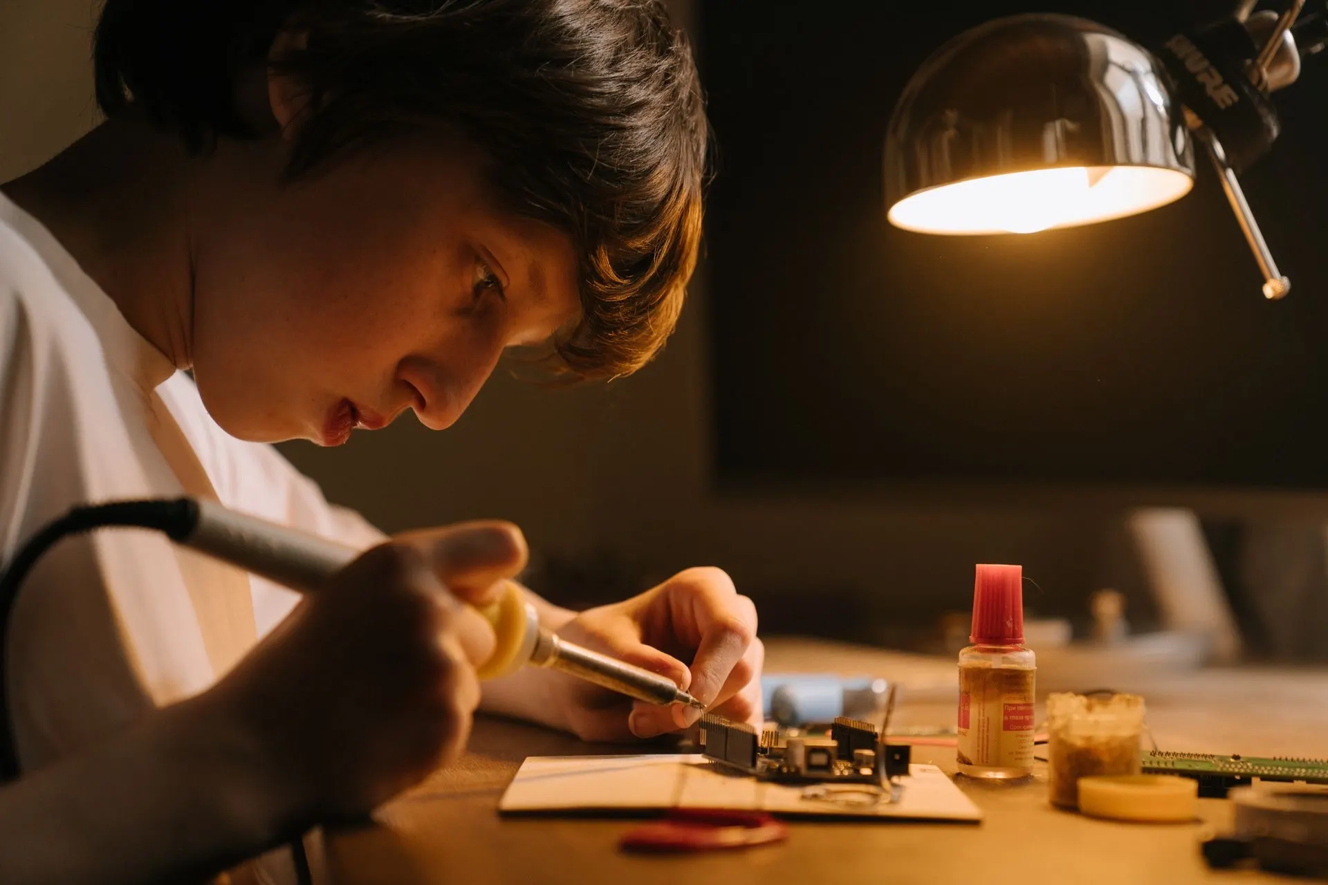 A teenager solders a circuit board under a desk lamp