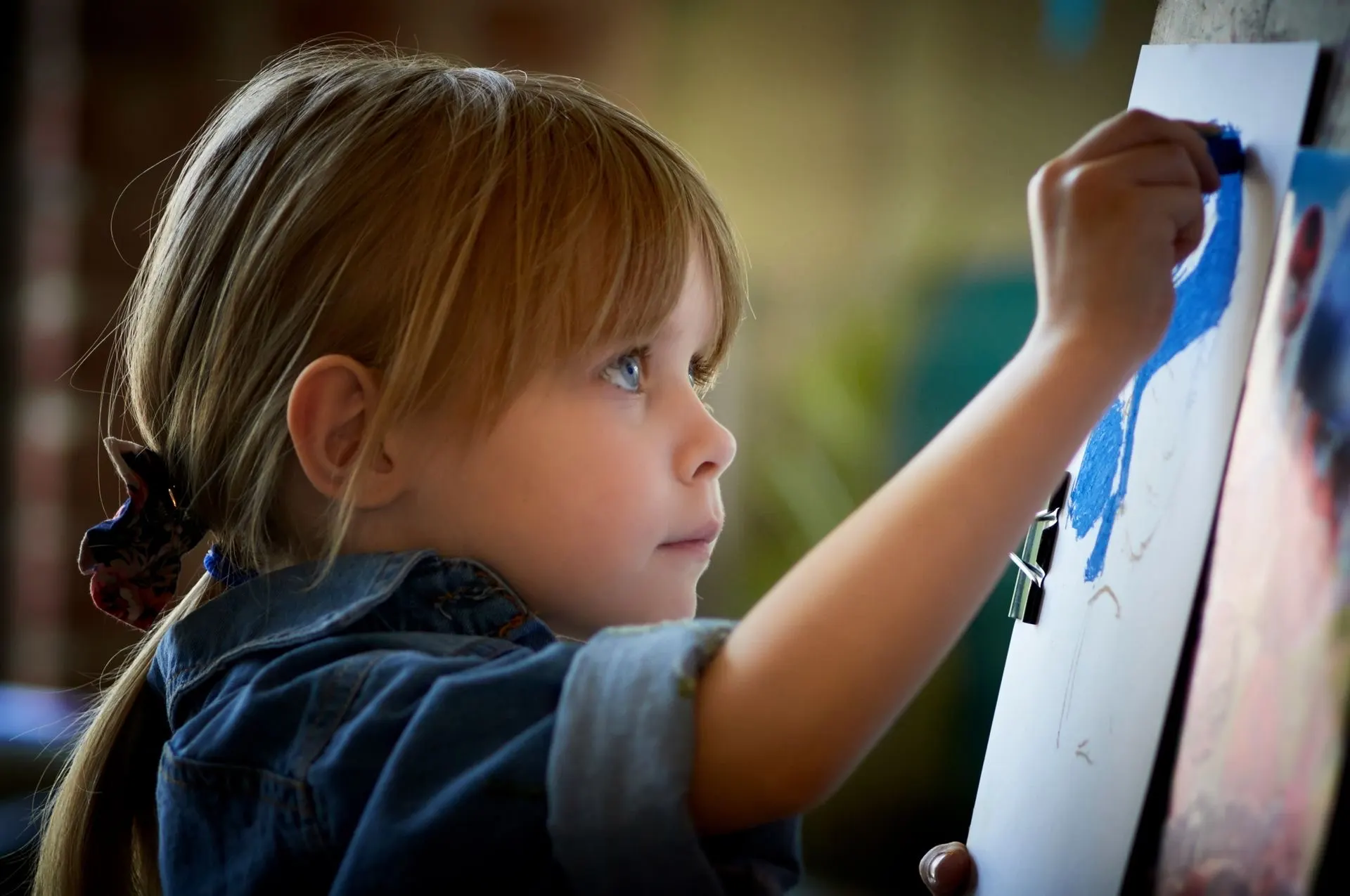 A young girl draws with a blue crayon at an easel, fully absorbed