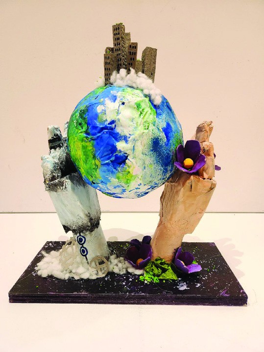 Photograph of a mixed media sculpture of two hands holding up a painted globe topped with cardboard skyscrapers