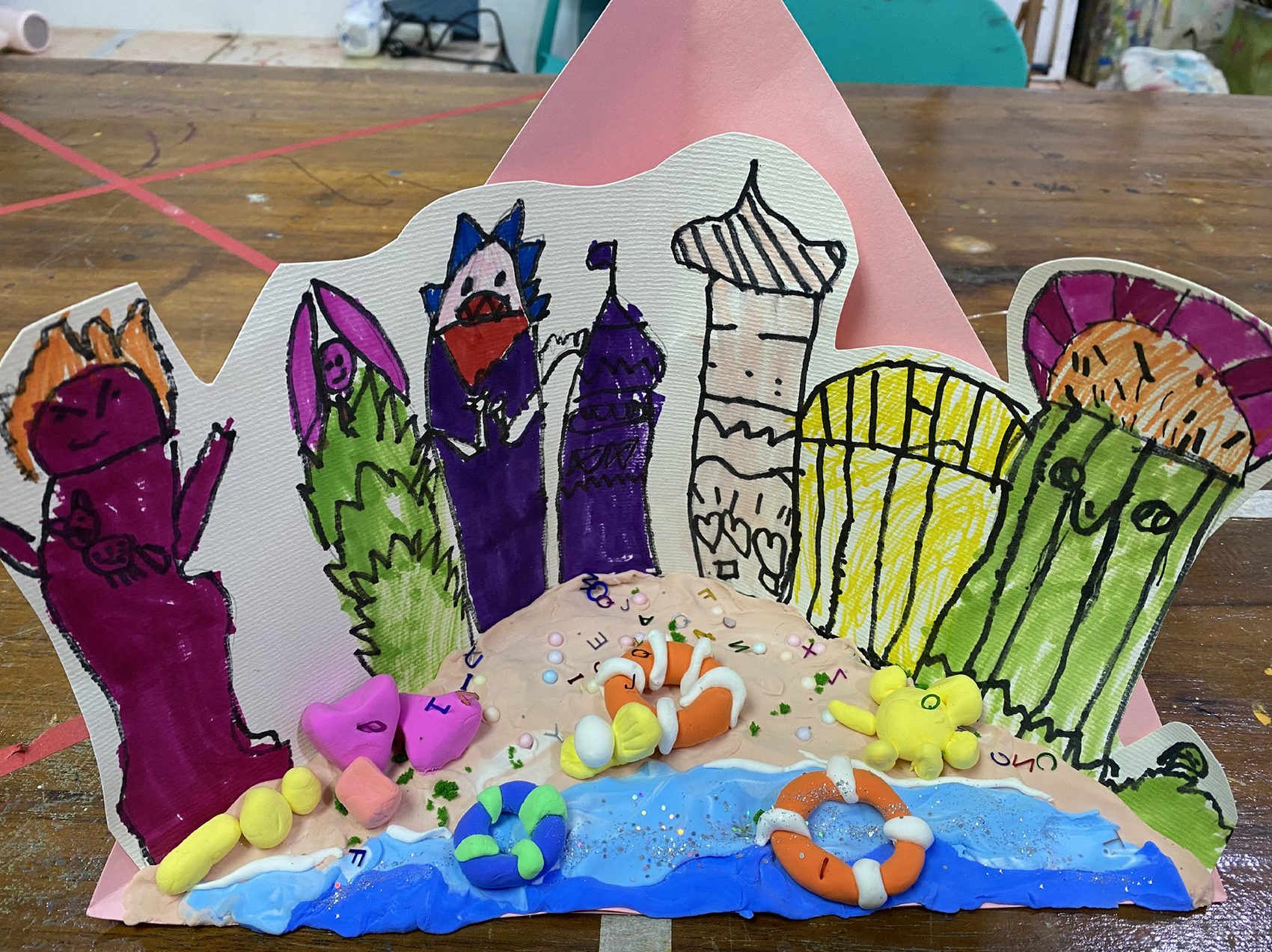 Paper-cut seaside town standing upright behind a modelled clay beach with lifebuoys and a glitter sea