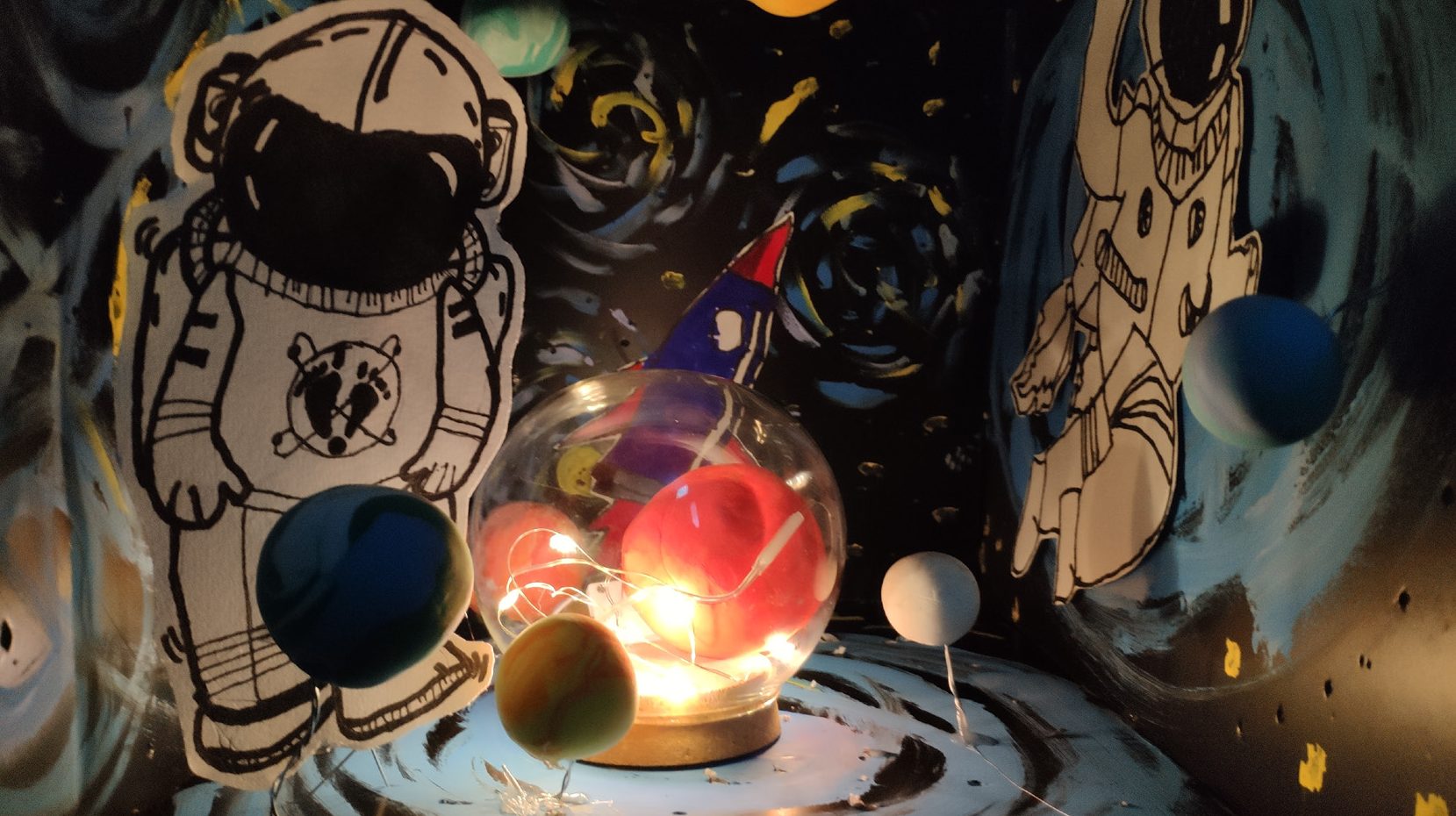 Cut-paper astronauts and clay planets arranged around a glowing glass-sphere lamp in a dark painted space scene