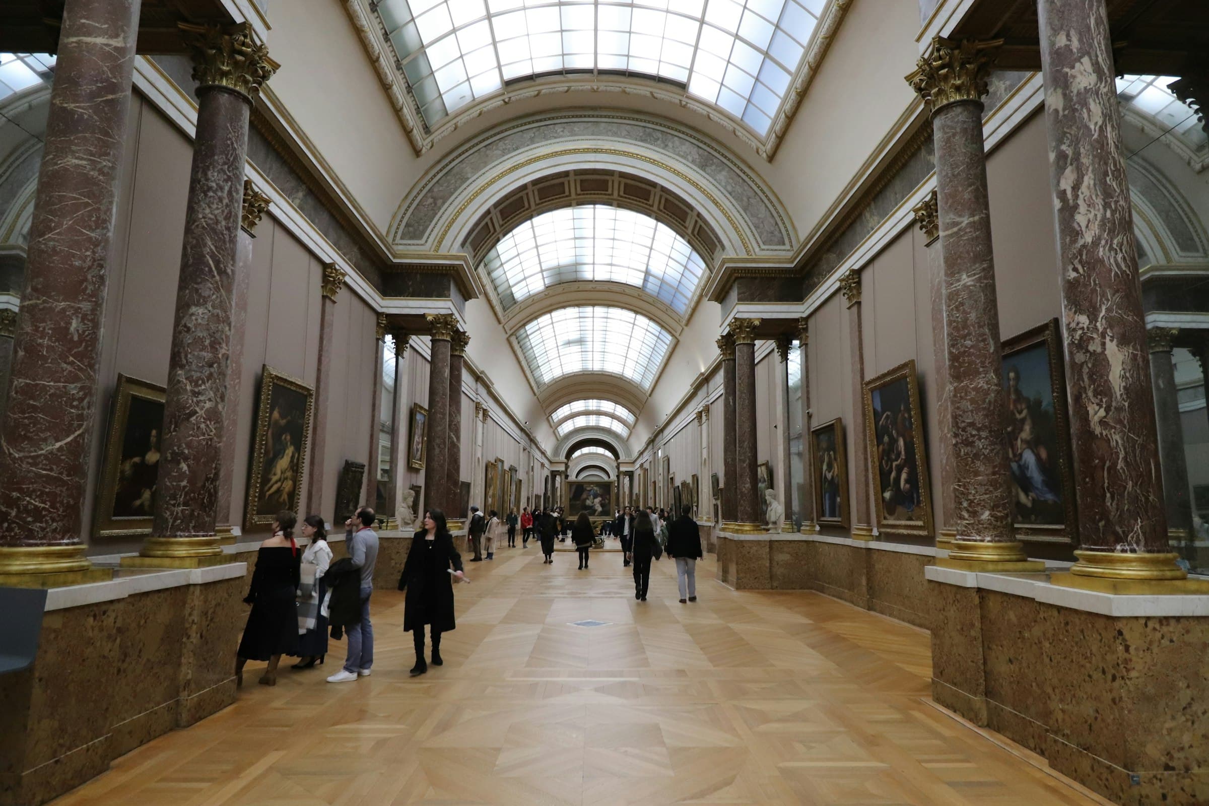 Visitors walking the skylit Grand Gallery of the Louvre past rows of gilt-framed paintings