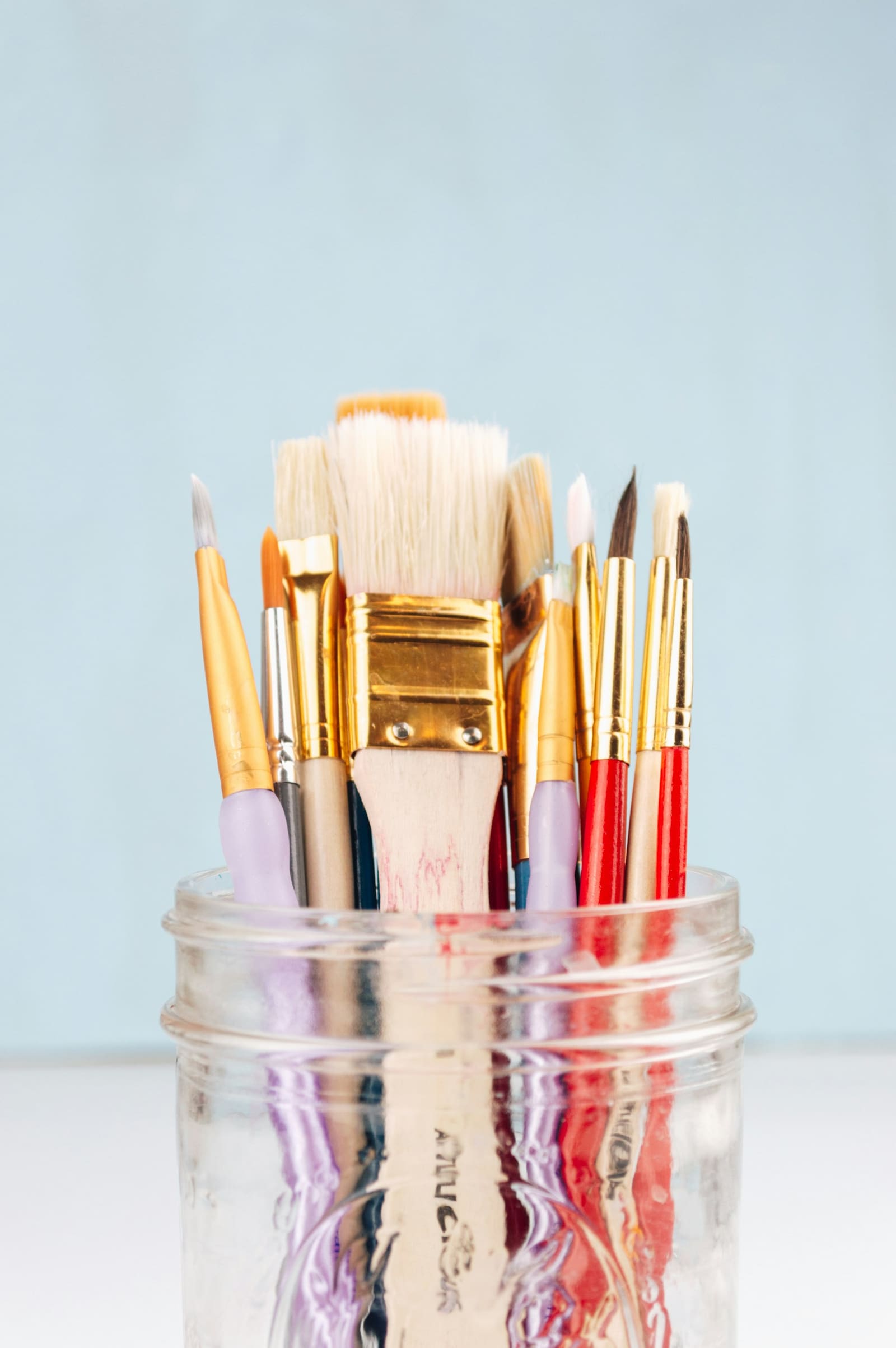 Assorted paintbrushes standing in a glass jar against a pale blue background