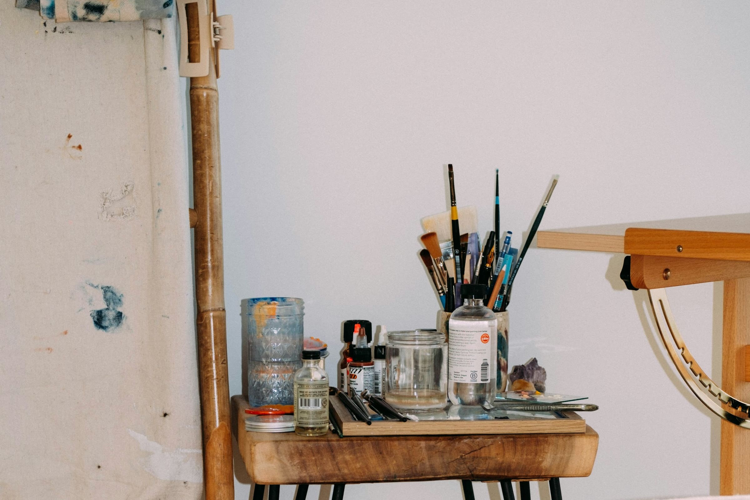Corner of a daylit teaching studio: brushes, inks and mixing jars set out on a timber side table beside a primed canvas