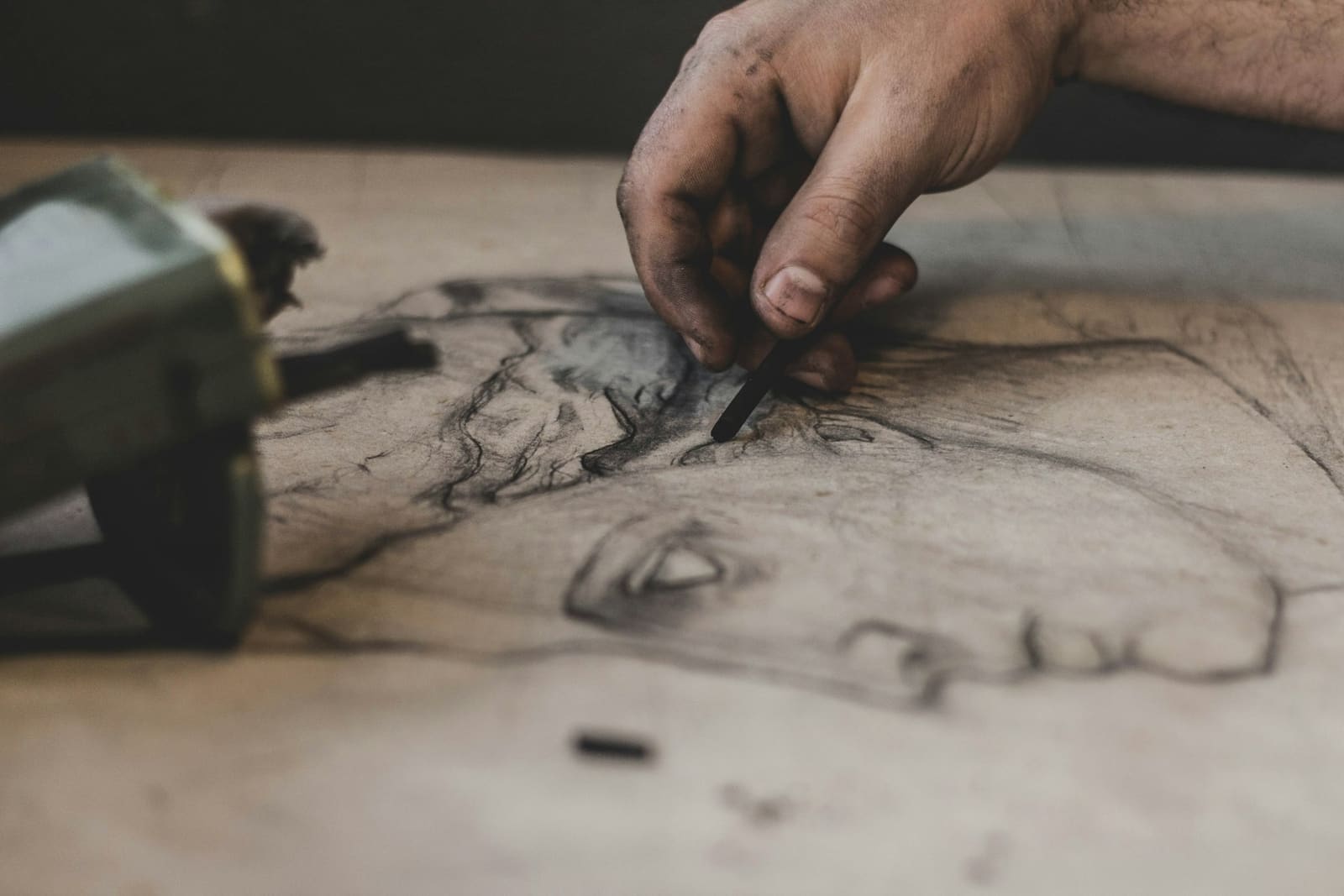 A charcoal-dusted hand shading a portrait study with a charcoal stick