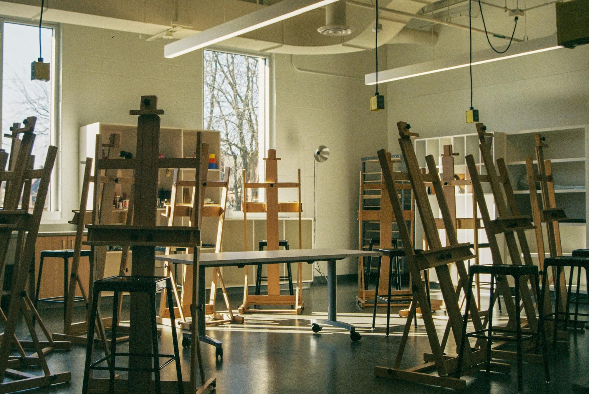 A sunlit studio classroom filled with wooden easels and stools around a worktable