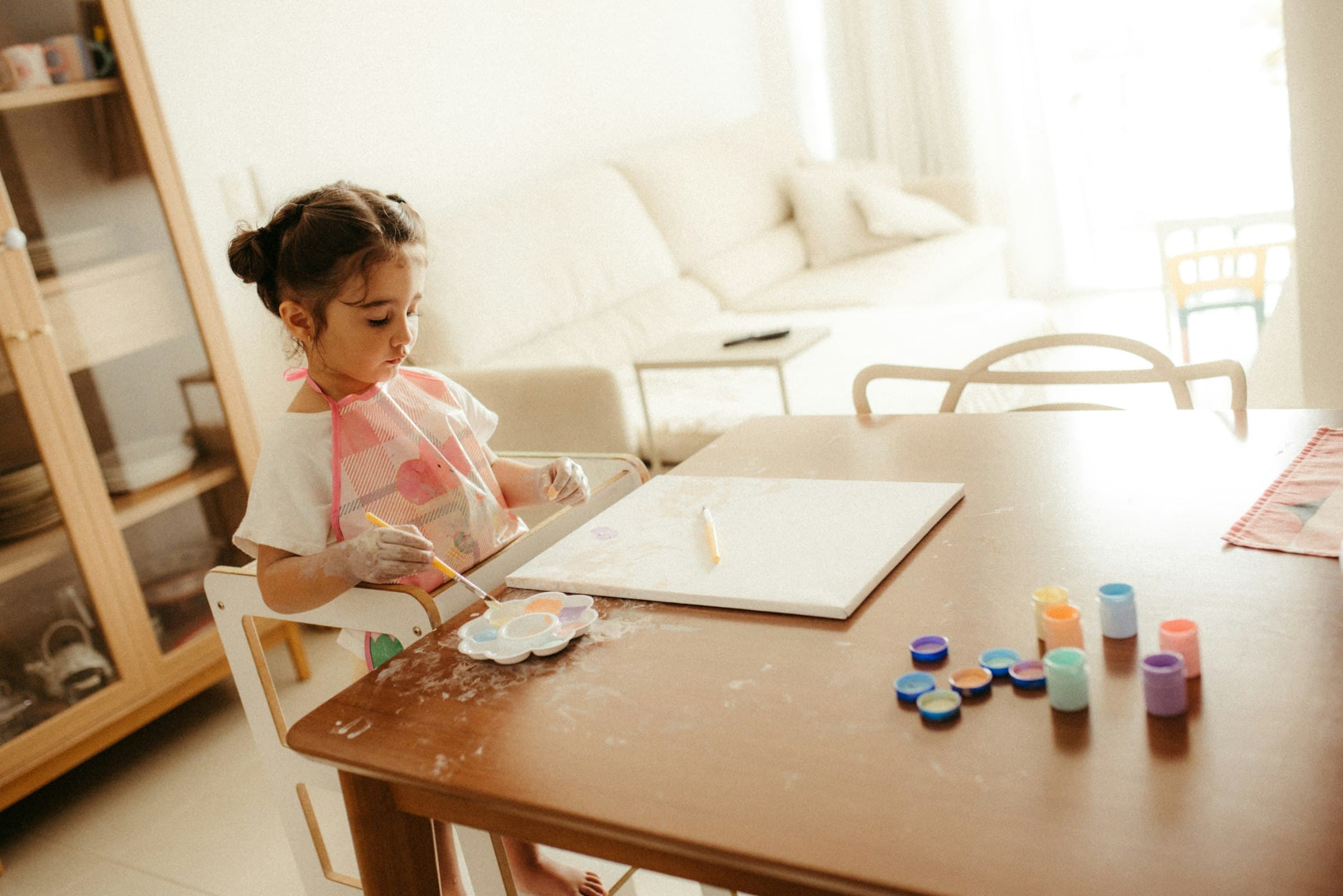 A young girl painting at a dining table in a sun-flooded living room