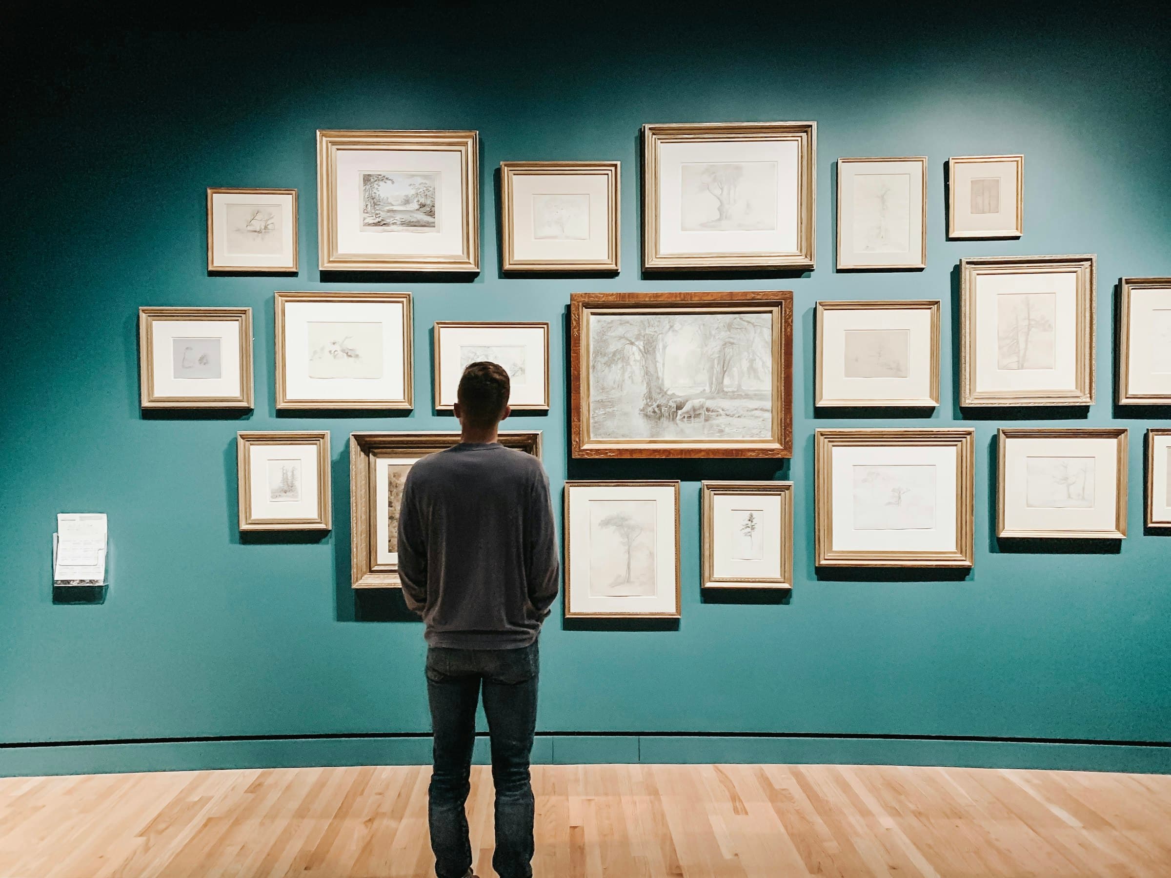 A visitor standing before a teal gallery wall hung with rows of gold-framed pencil drawings