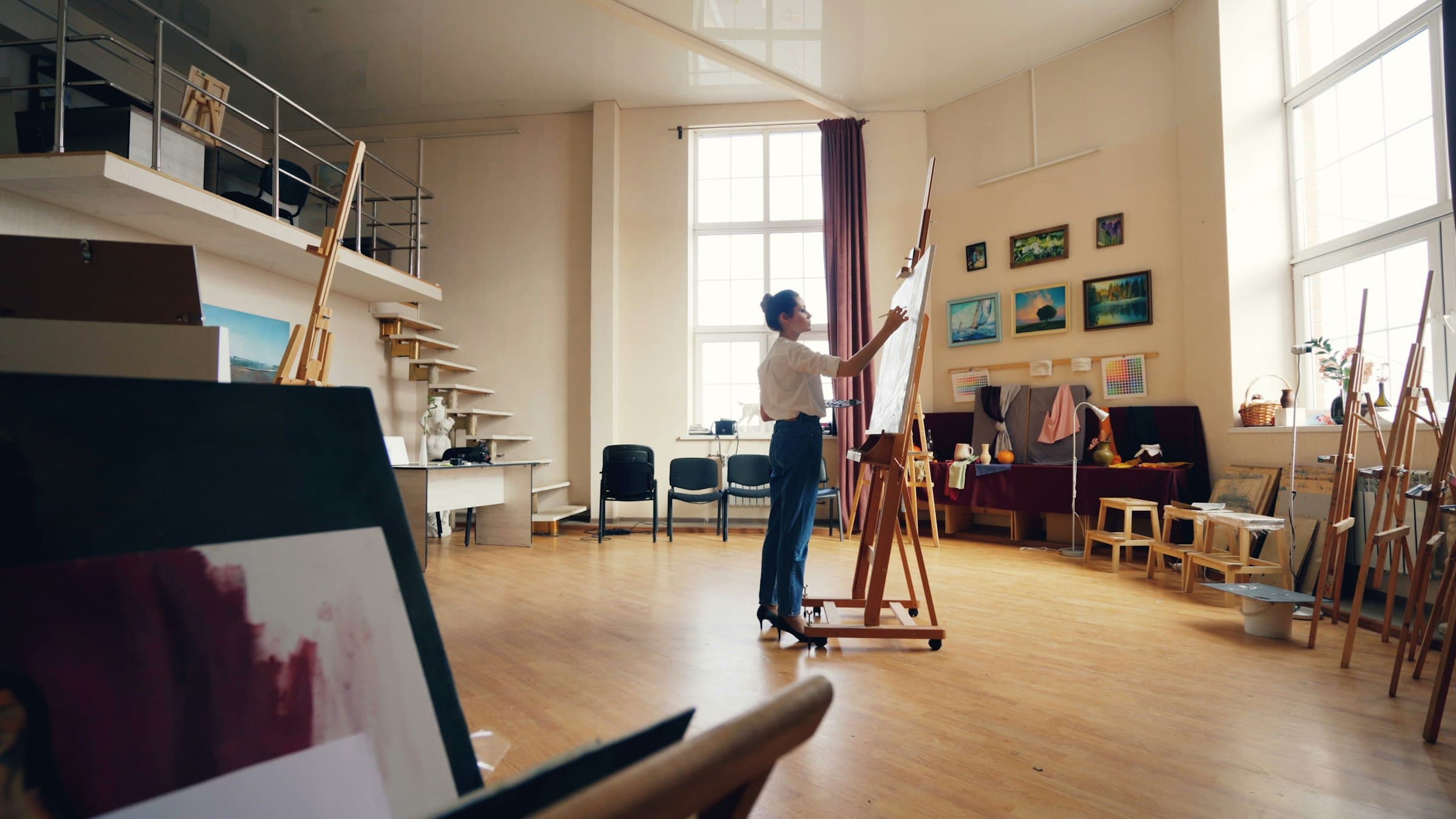 An artist painting at a full-size easel in a double-height loft studio hung with framed landscapes