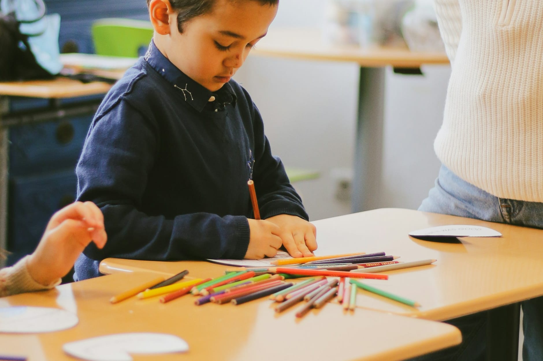 Child drawing at a light-filled studio table spread with coloured pencils while a teacher stands alongside