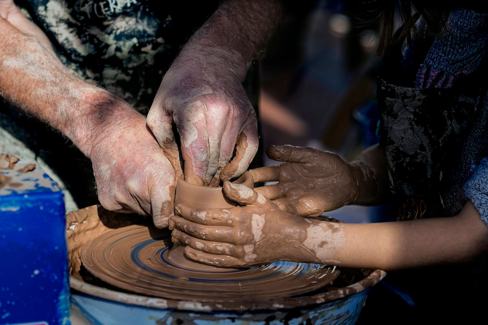 Adult hands guiding a child's clay-covered hands as they shape a pot on a potter's wheel