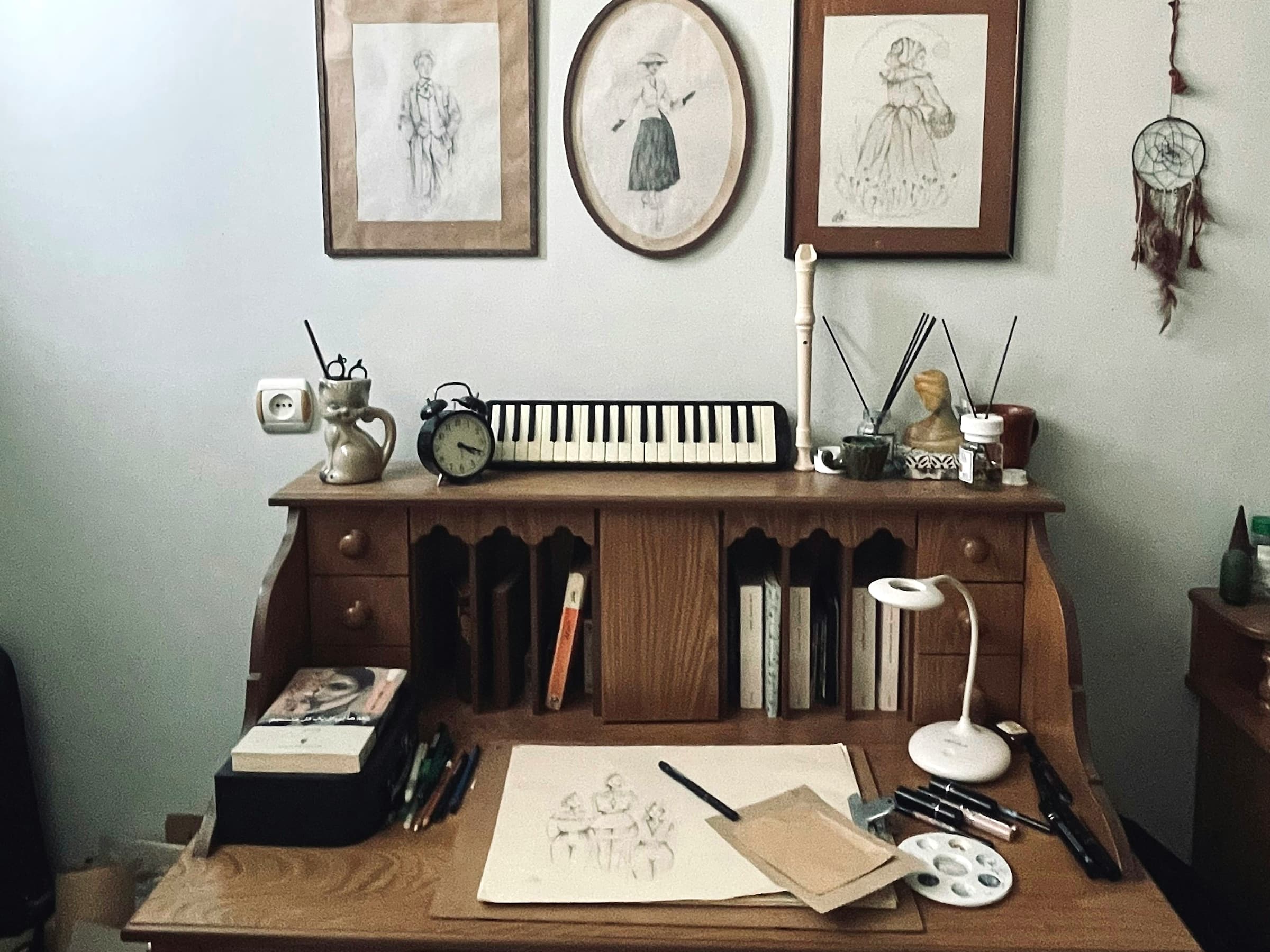 Home art desk with a half-finished pencil sketch, palette and pencils, framed drawings hung on the wall above