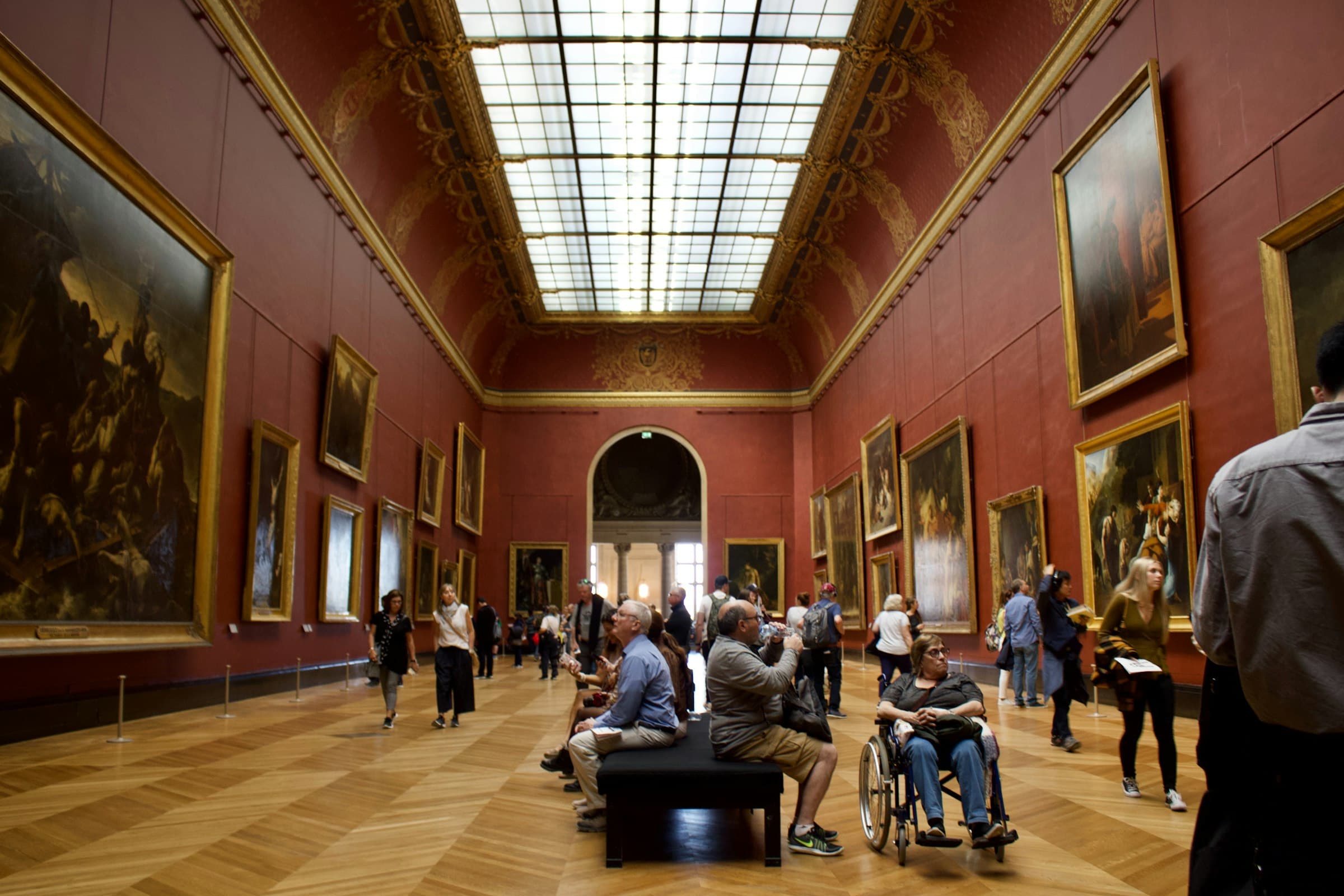 Visitors studying rows of gold-framed old master paintings in a tall museum hall with deep red walls and a glass skylight, some seated on a bench looking up at the canvases