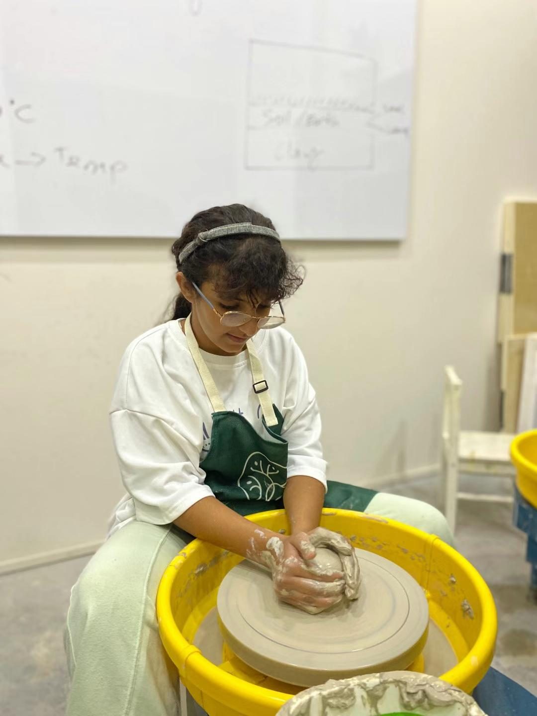 Inside a Tree Art studio — a student shaping clay on a pottery wheel, with pottery notes on the whiteboard behind her