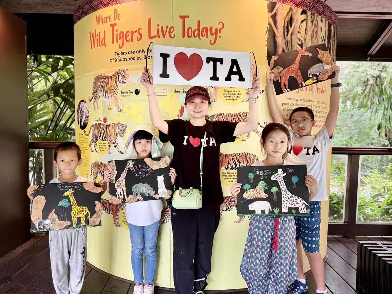 A small group at the zoo's tiger exhibit holding up animal collage artworks and an I-love-TA banner