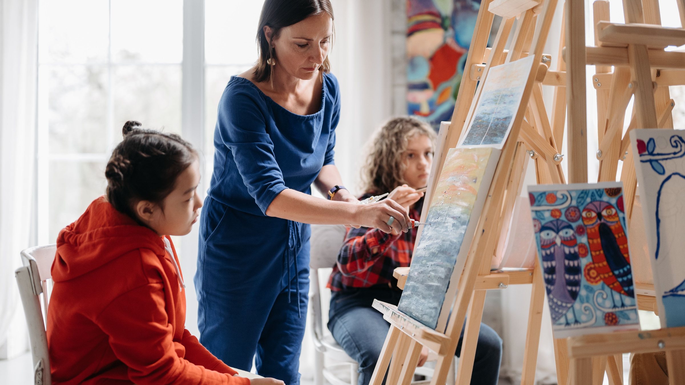 Art teacher guiding young students painting at easels in a bright studio