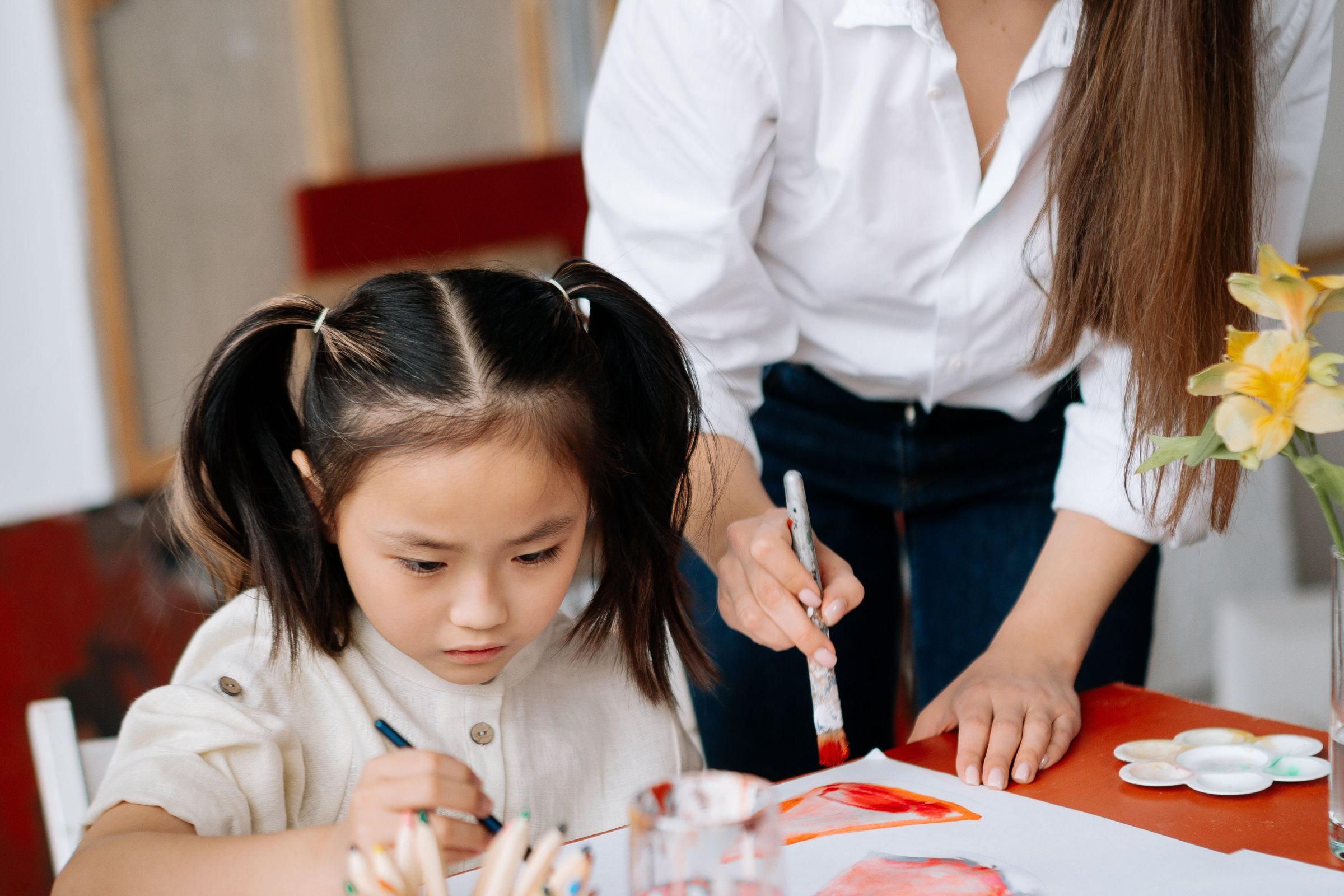 Teacher's hand guiding a young girl as she paints at her table in an art class