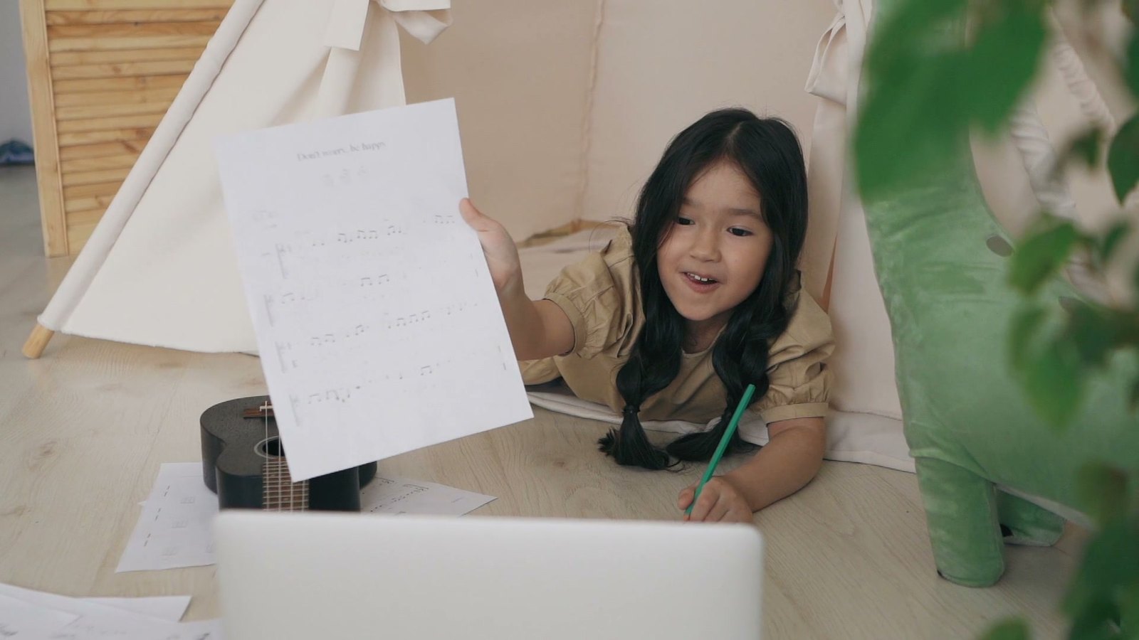 Girl in a play tent holding her drawing up to the open laptop during an online lesson