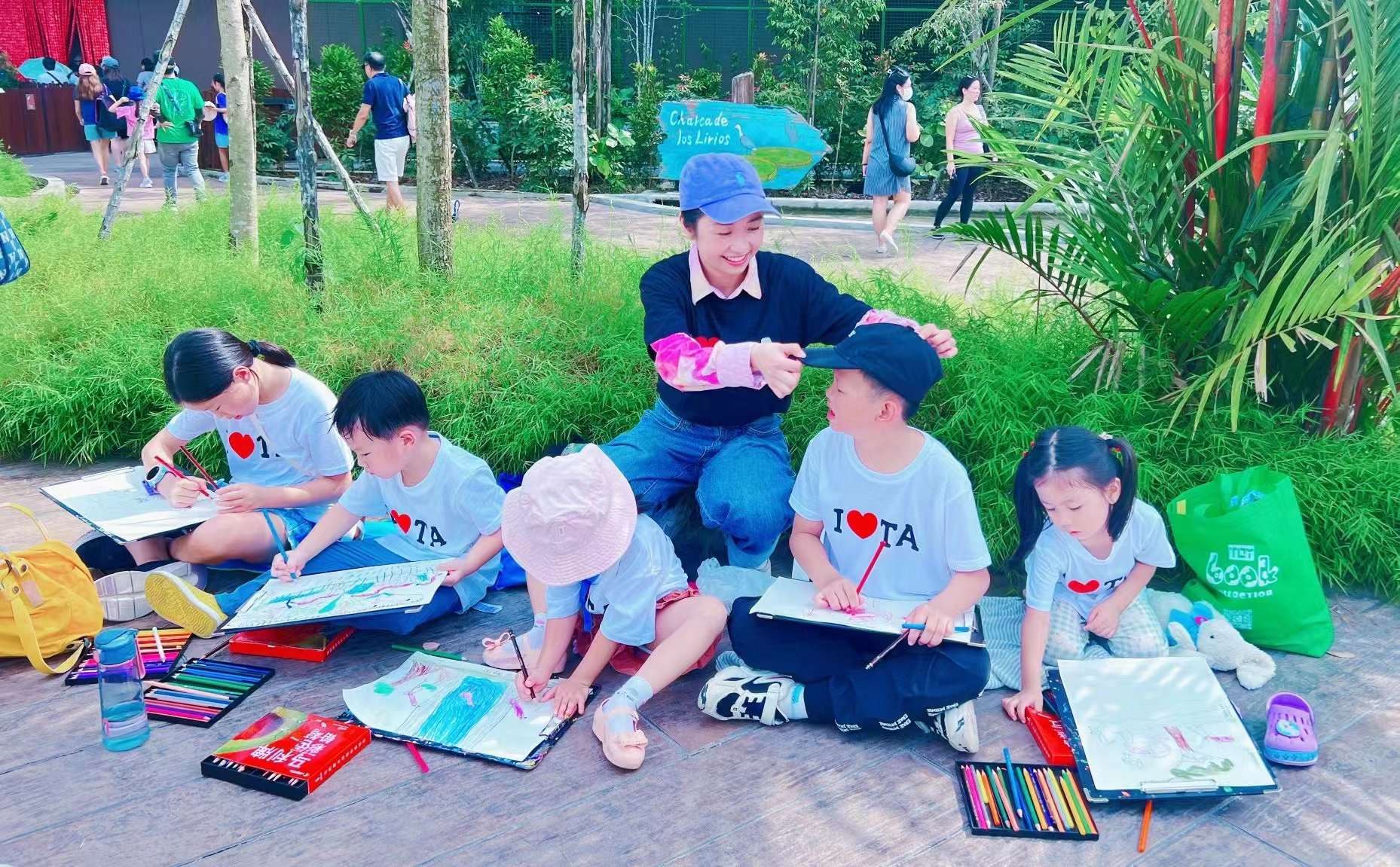 TREE ART teacher kneeling among children in I-love-TA tees sketching on drawing boards in a lush park
