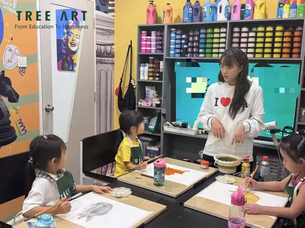 TREE ART teacher demonstrating a painting technique to three young girls in aprons at their studio table